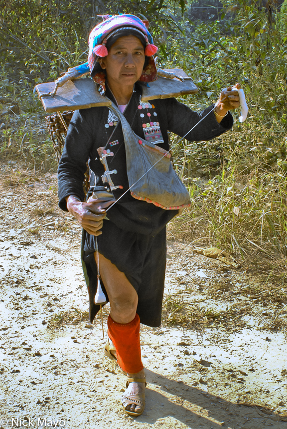 A Hani woman from Da Fung Liang walking home carrying a backstrap basket attached to a wooden yoke while spinning cotton on a...