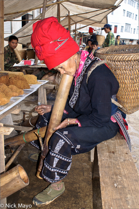 A Red Yao woman carrying a basket and wearing traditional clothes and hat smoking tobacco through a bamboo pipe in the market...