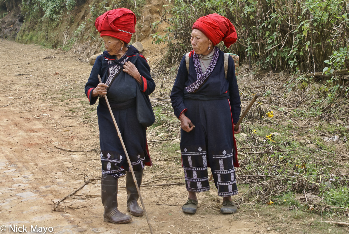 Two older Red Yao women wearing traditional clothes and hats walking home from Maandi market.