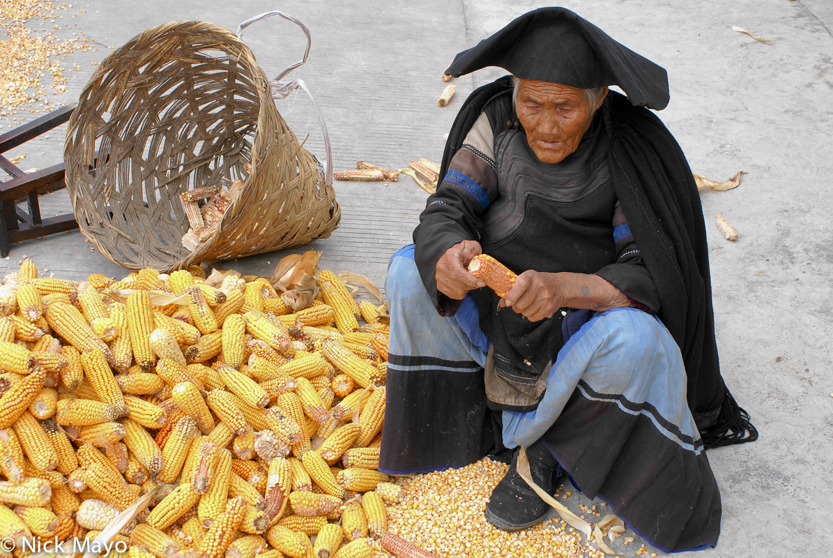 A Yi woman in a traditional cape and hat shucking corn cobs at Tu Wu village with a wicker container to hold the husks.