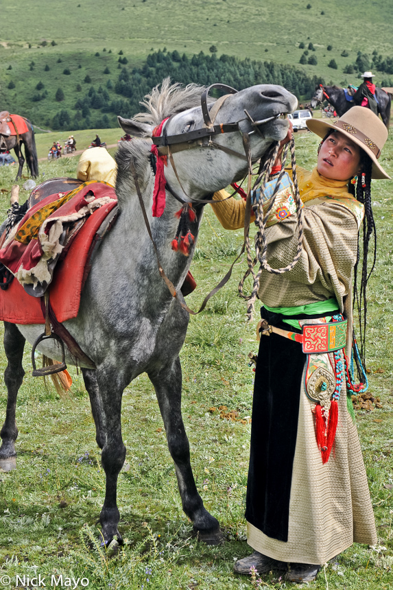 A young Tibetan woman with braided hair and a bagu hanging from her waist band about to leave by horse from a monastery festival...