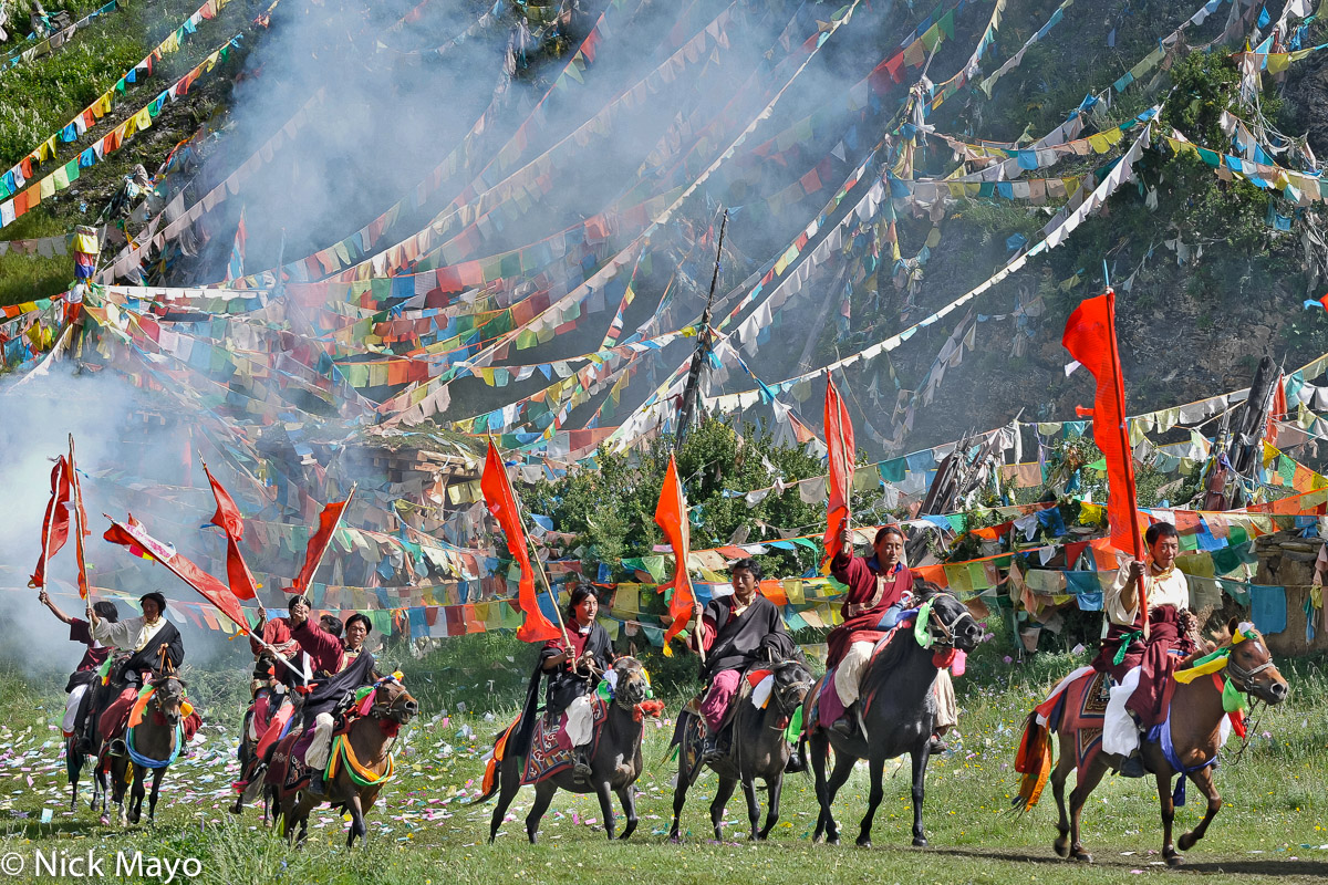 Standard bearing Tibetan horsemen passing prayer flags at the entrance to a grassland festival near Manigango.