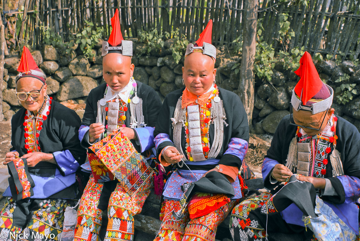 Four Hongte Yao women wearing traditional clothes, breastpieces and peaked hats stitching in Ping An Jie.