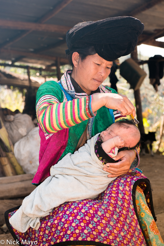 A White Hmong woman wearing a traditional peaked turban shaving her child's head in the village of Ju Guo Yue.