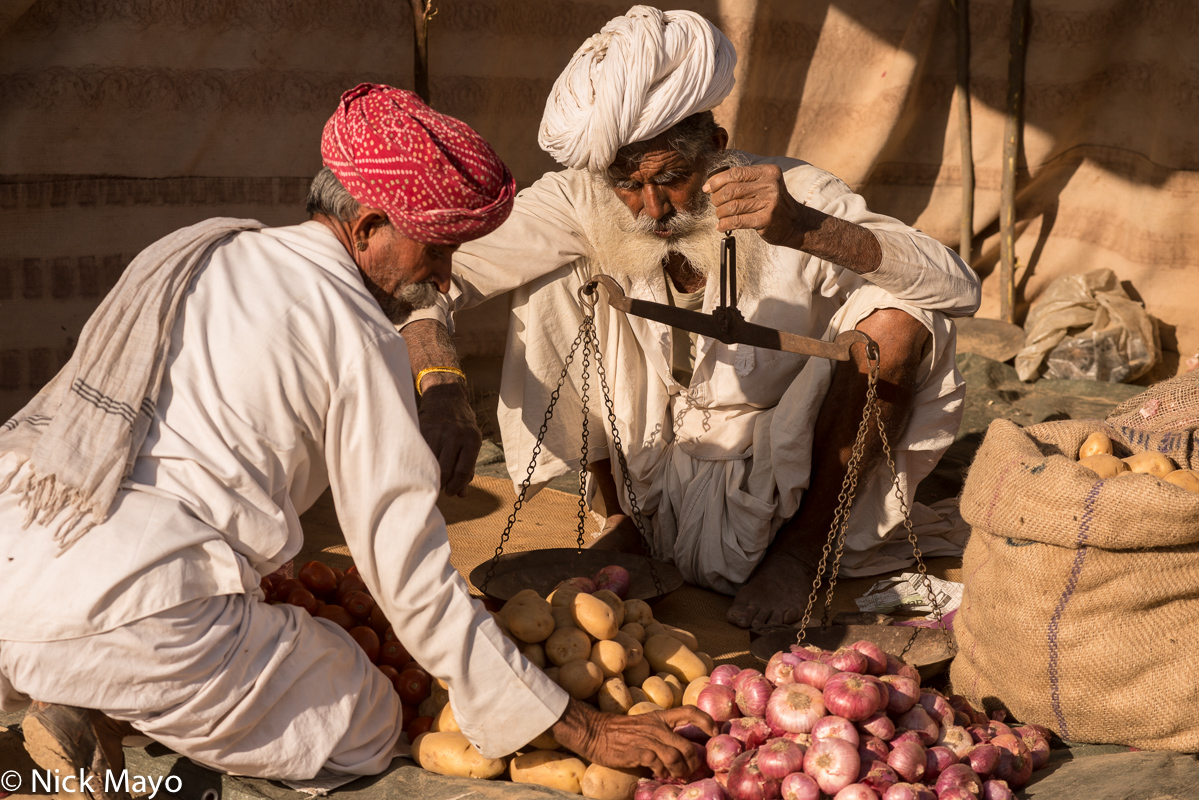 A vendor using a hand held weighing scale to measure the weight of onions and potatoes being bought by a Rabari man during the...