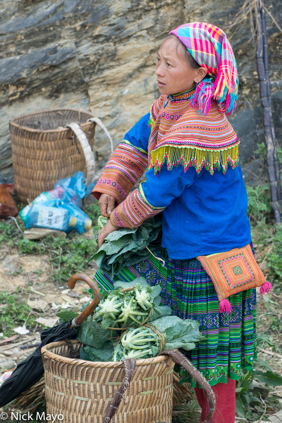 A Hmong woman, wearing a pleated skirt and checked headscarf, with a basket of vegetables at Si Ma Cai market.