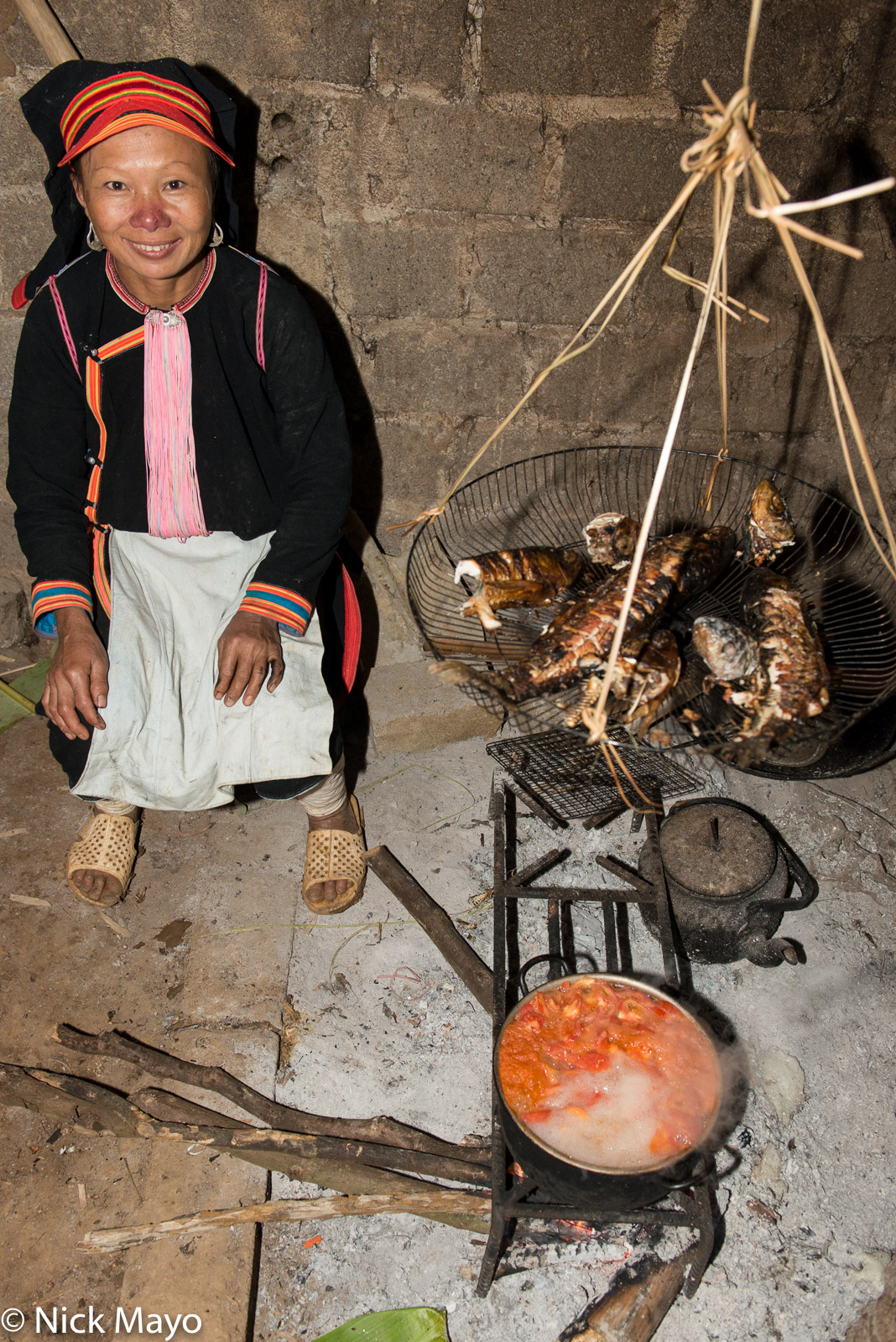 A Black Dao (Yao) woman wearing traditional dress of hat, earrings and apron cooking fish and soup in Ban Then Sin.