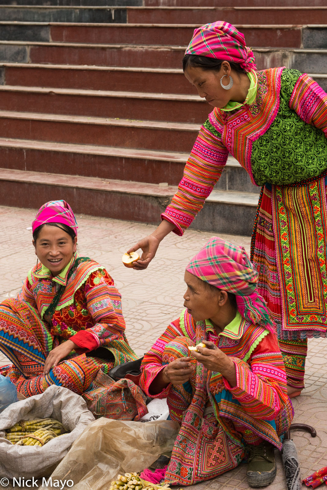 Three Flowery Hmong woman in traditional clothes, one wearing moon earrings, selling vegetables at Su Phi market.