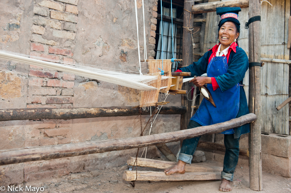 A Bai Yue Hani woman, in traditional clothes and hat, weaving at a foot treadle loom in the village of Yi Shan Chuan.