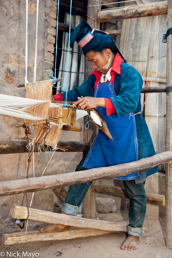 A Bai Yue Hani woman, in traditional clothes, hat and earrings, weaving at a foot treadle loom in the village of Yi Shan Chuan...