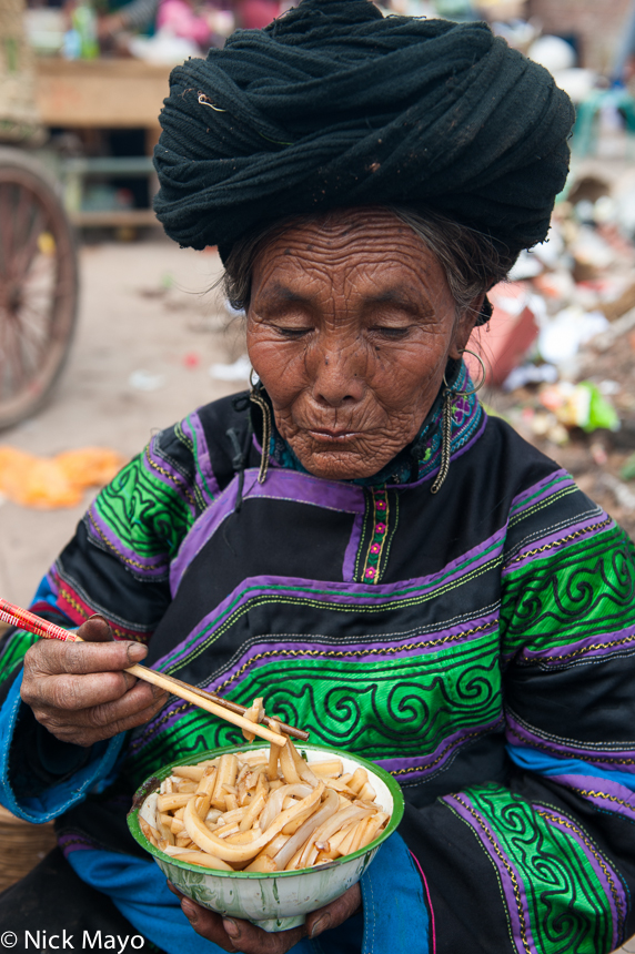 A Yi women dressed in traditional clothes, turban and earrings eating a lunch of noodles at Shau Mu Shan market in Daliangshan...