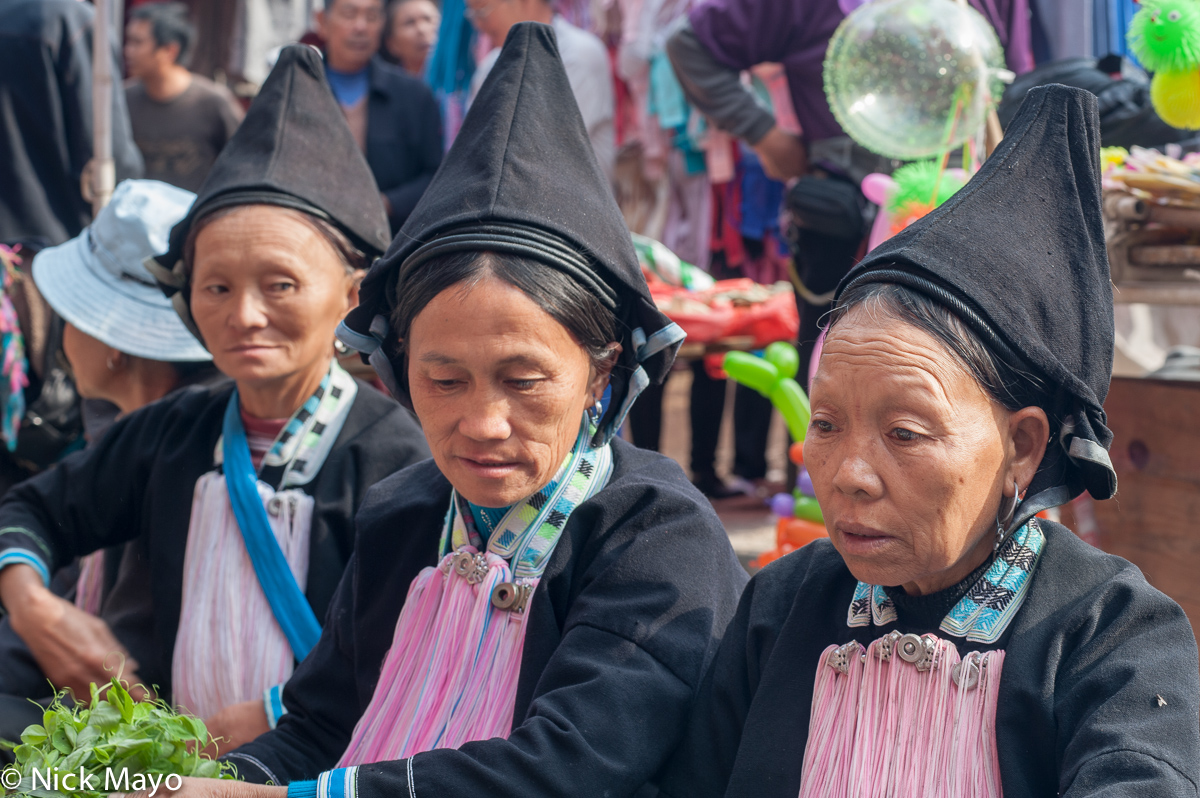 Three Yao women from the Meng Chiao area, in peaked black hats, selling vegetables at market.