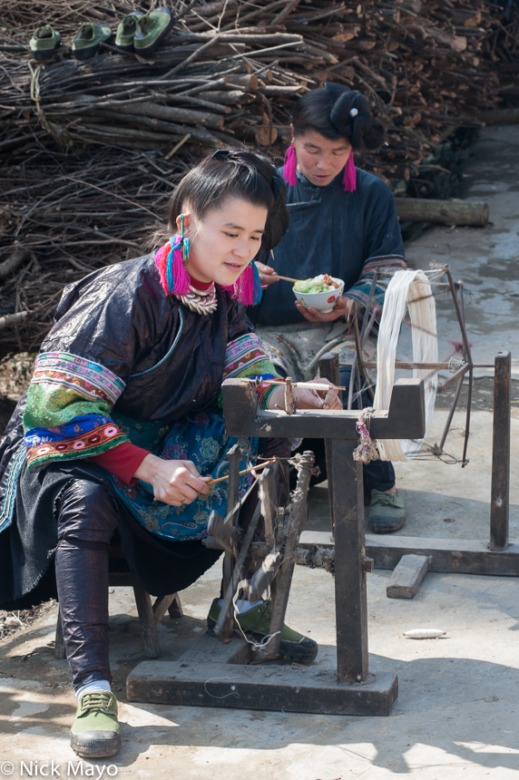 A young woman from Miao Peng spinning thread on a hand operated spindle while her mother eats lunch.