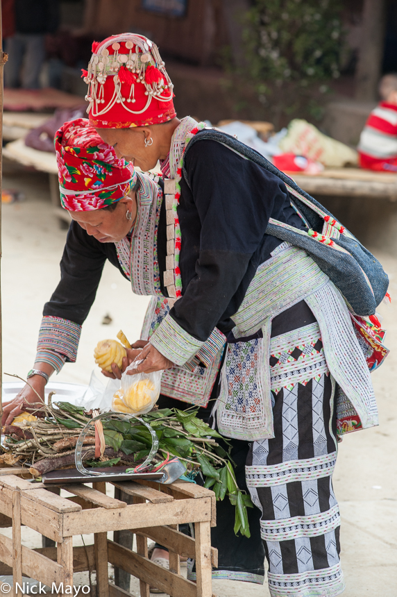 Two Red Dao (Yao) women, in their traditional dress, earrings and hats, shopping for pineapples at Muong Hum on market day.