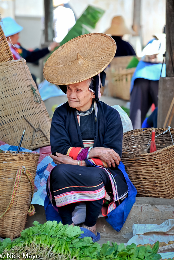 A Hua Yao Dai woman, wearing her traditional clothes and hat, selling vegetables at Mosha market surrounded by baskets.