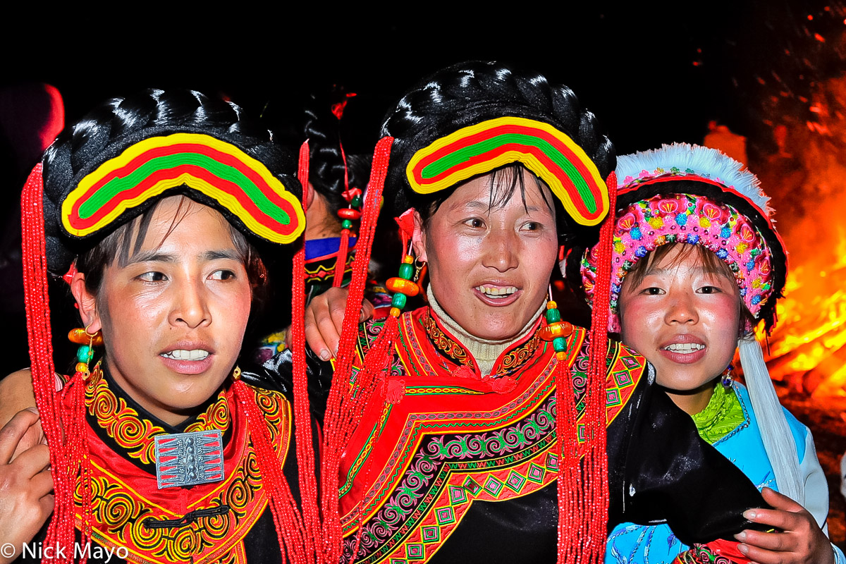 Three Yi women, two wearing fake hairpieces and beaded earrings and the third a traditional hat, by a bonfire at a festival in...