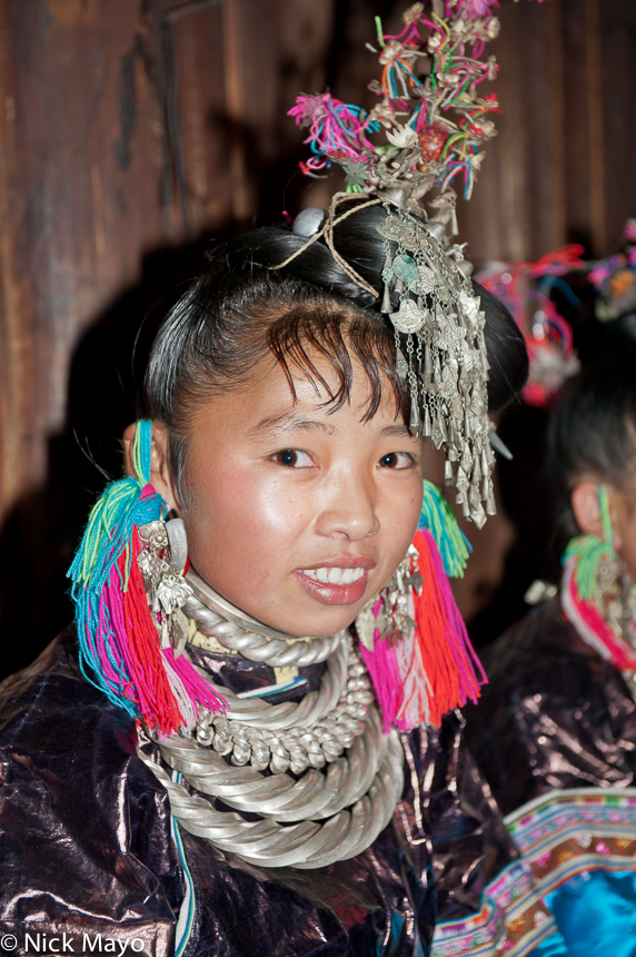 A Miao bridesmaid wearing a silver hair piece, heavy necklaces and multicoloured tasselled woollen earring at a wedding in the...