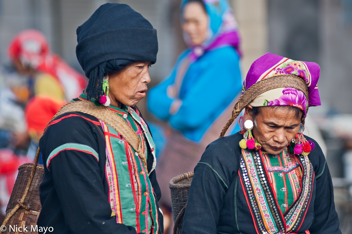 Two traditionally dressed Bulang women, with turbans and pom pom earrings, carrying their baskets in Mengzhe market.