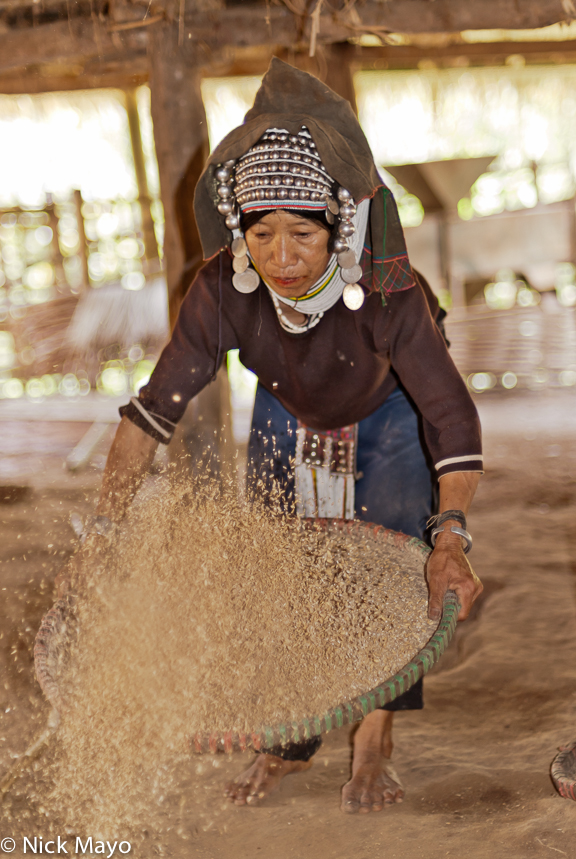 An Akha (Hani) woman, wearing a traditional headdress, winnowing paddy rice beneath her house in Ba Bei.