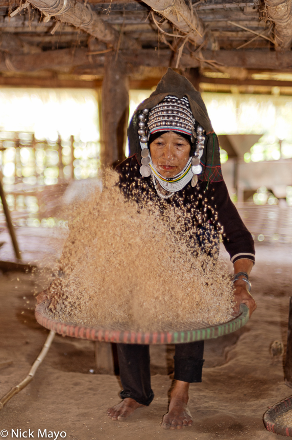 An Akha (Hani) woman, wearing a traditional headdress, winnowing paddy rice underneath her house in Ba Bei.