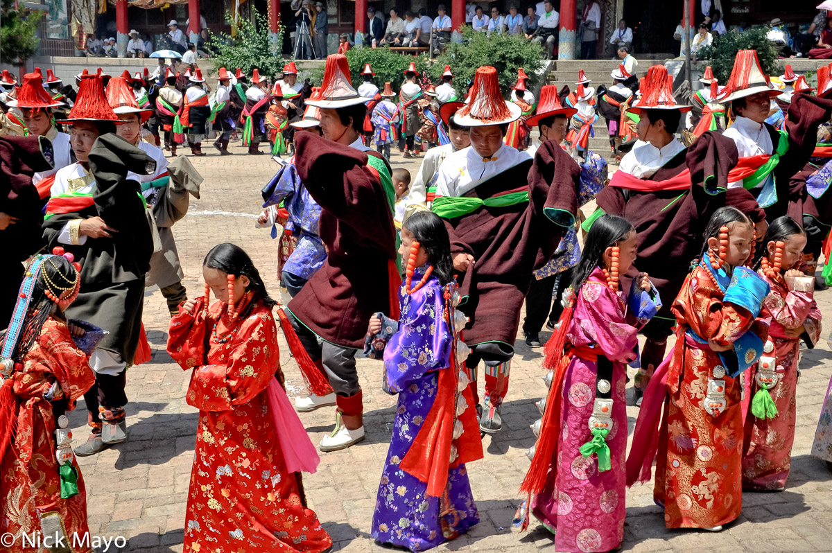 Tibetan men, wearing red tasselled hats, and young girls, wearing heavy beaded hair pieces, dancing at a monastery festival in...