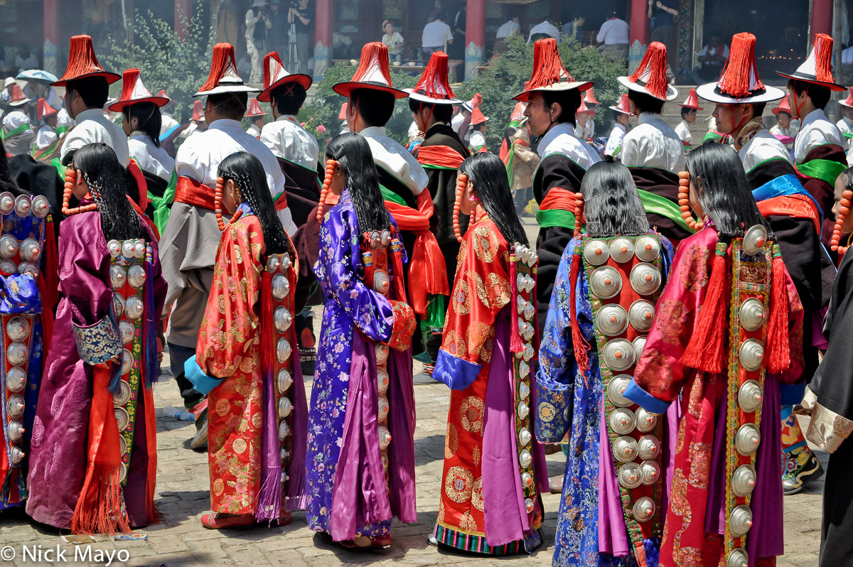 Tibetan men, wearing red tasselled hats, and girls, with heavy studded hair pieces hung from their braided hair, in a procession...