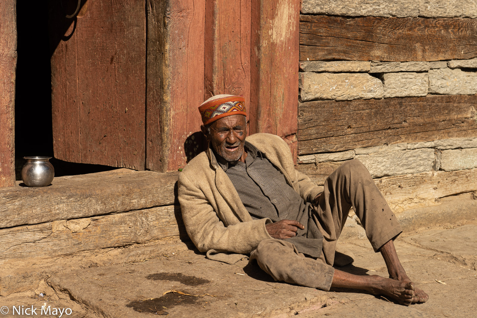 A Hallan-I man relaxing outside his traditional Kulu house built with layered wood and stone walls.